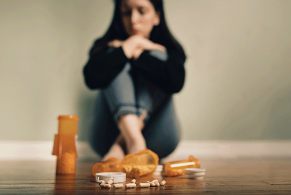 an upset woman sits on the ground in front of empty pill bottles on the floor and asks herself what are benzos