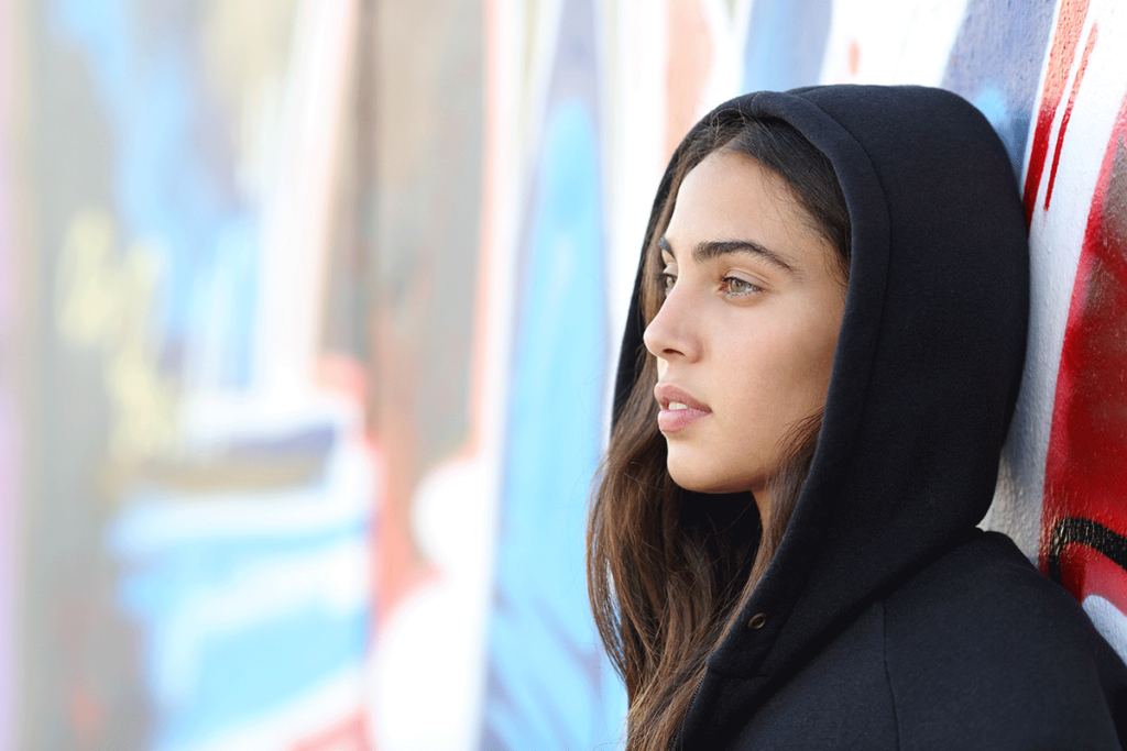young woman in hoodie seated against a colorful wall outside contemplating the four signs of cocaine addiction you should know