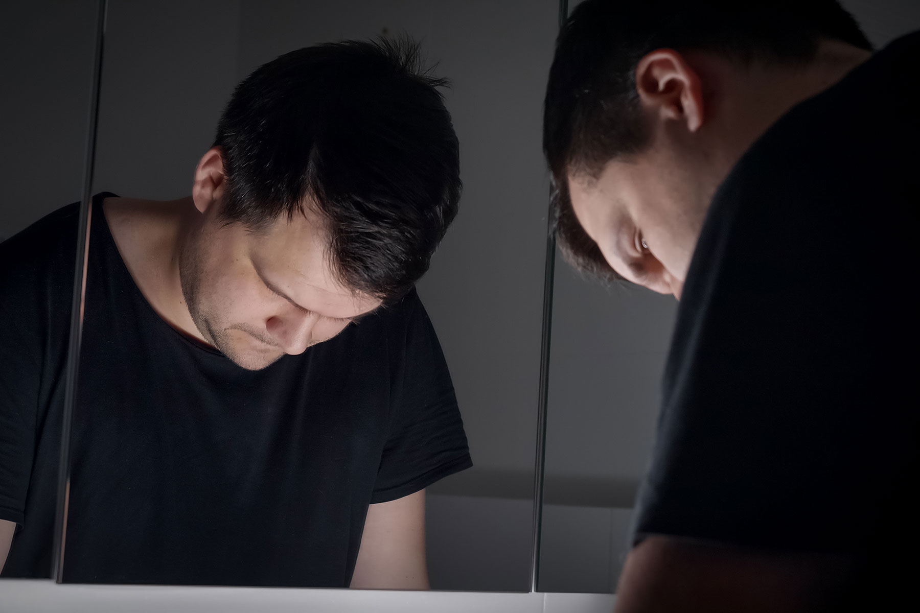 young man standing in front of bathroom mirror with head bowed reflecting on the 5 causes of substance abuse