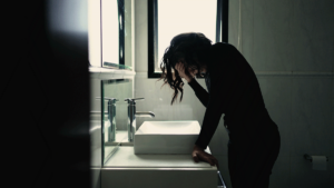 young woman hunched over bathroom sink holding her head in her hands experiencing the dangers of mixing adderall and alcohol