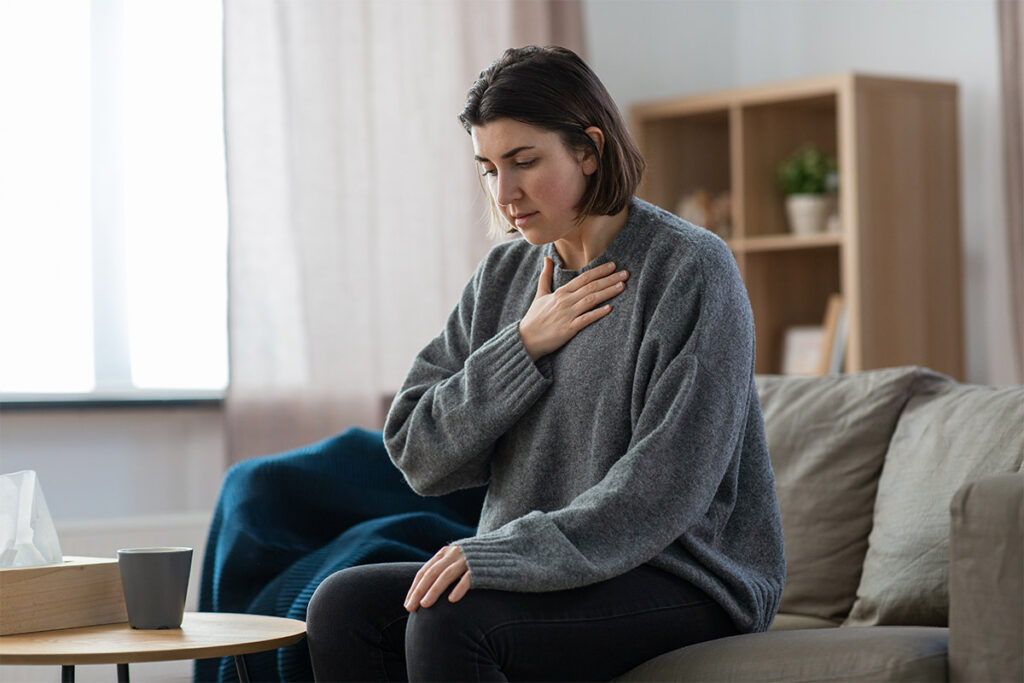 young woman seated on her couch at home clutching her chest learning how to recognize and understand panic attacks vs. anxiety attacks.