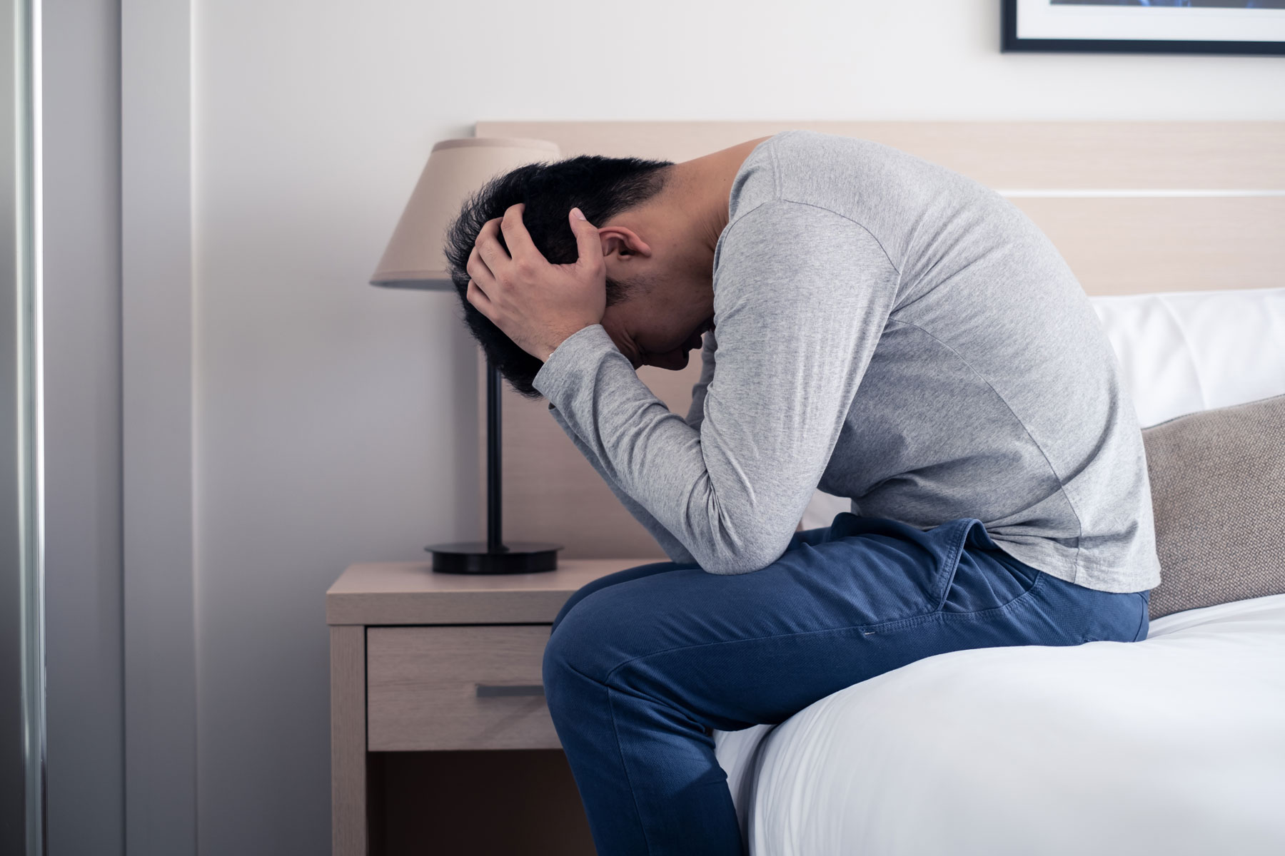 A man sits on the edge of his bed with his head in his hands. He's dealing with memory loss and mood swings, both symptoms of alcohol-induced dementia.