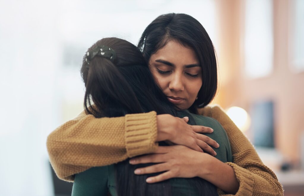 Two women hugging each other closely, showing empathy and comfort, with one woman’s eyes closed as they share an emotional moment.