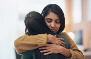Two women hugging each other closely, showing empathy and comfort, with one woman’s eyes closed as they share an emotional moment.
