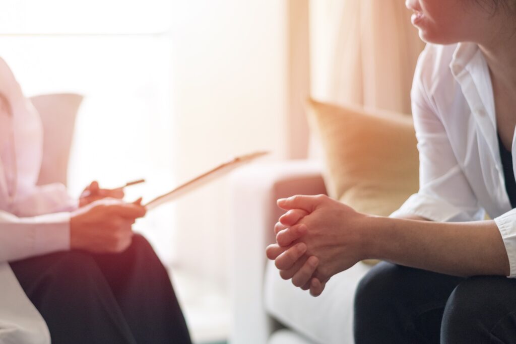 Close-up of a therapy session showing a person sitting with clasped hands, speaking to a counselor holding a clipboard and pen in a softly lit room.