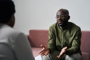 A man in glasses and a green shirt sits on a couch, speaking openly to someone during a counseling session.