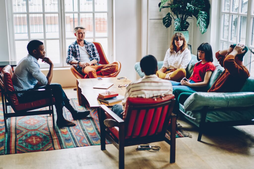 A group of people sits in a circle in a bright, cozy room, engaged in conversation. They sit on various chairs and sofas around a low coffee table with books and papers on it. Large windows and plants add natural light to the relaxed group setting.